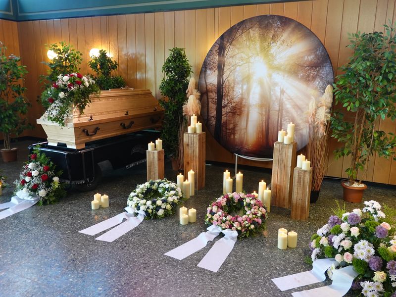 Holzsarg mit Blumenschmuck, Kränzen und Kerzen vor einem Lichtmotiv in der Trauerhalle