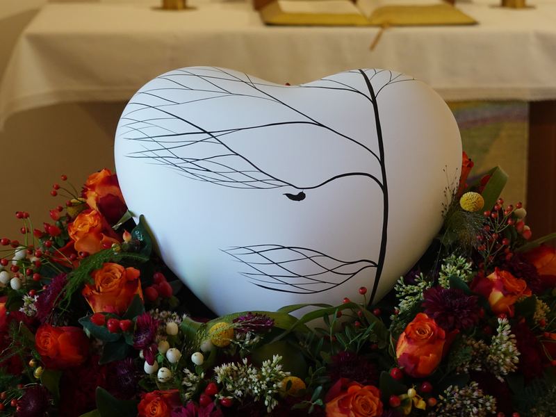 Weiße Urne in Herzform mit Zweigmotiv auf rotem Blumenkranz